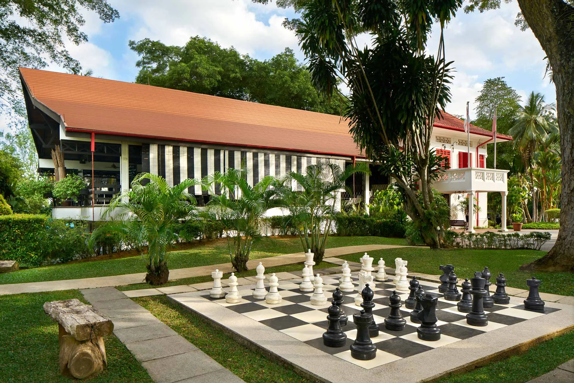 Outdoor giant chess set on a black-and-white chequered court with tropical gardens, a red-roofed clubhouse, and a colonial-style building in the background.