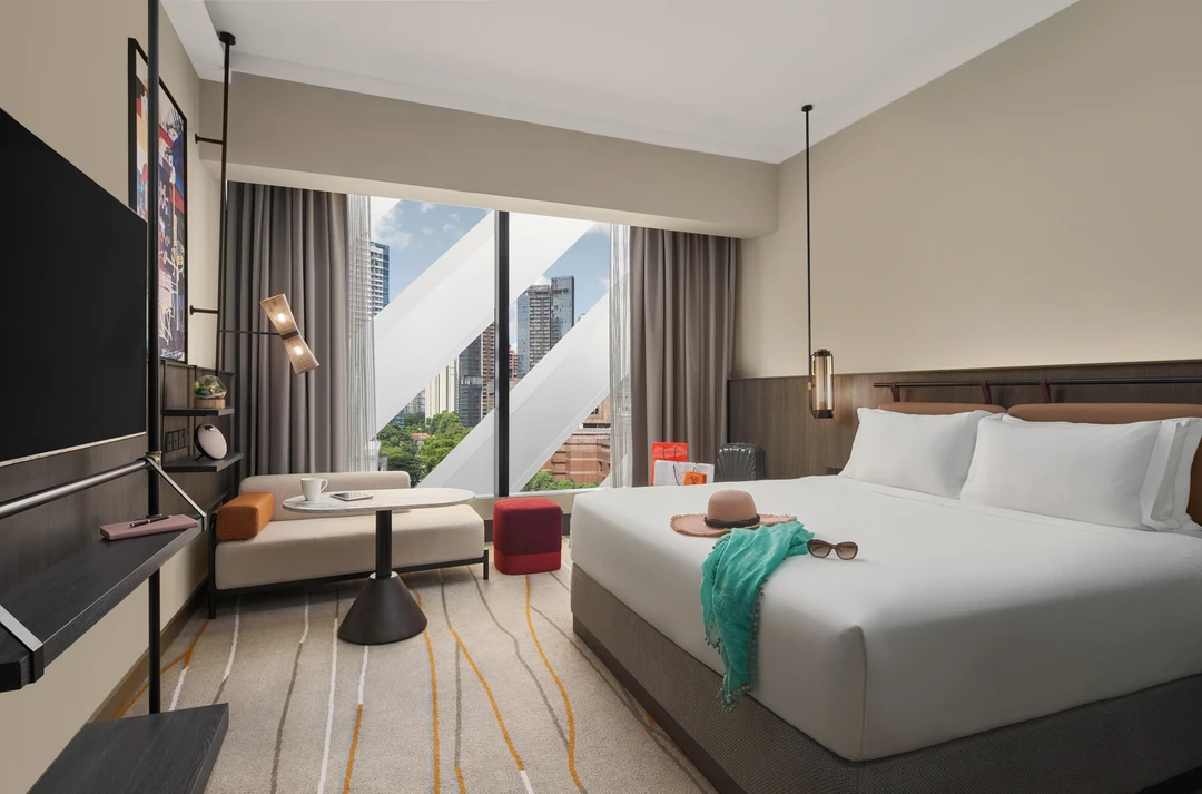 Modern hotel room with king bed dressed in white linen, a straw hat and turquoise scarf on the bed, floor-to-ceiling city-view window, and contemporary furnishings.