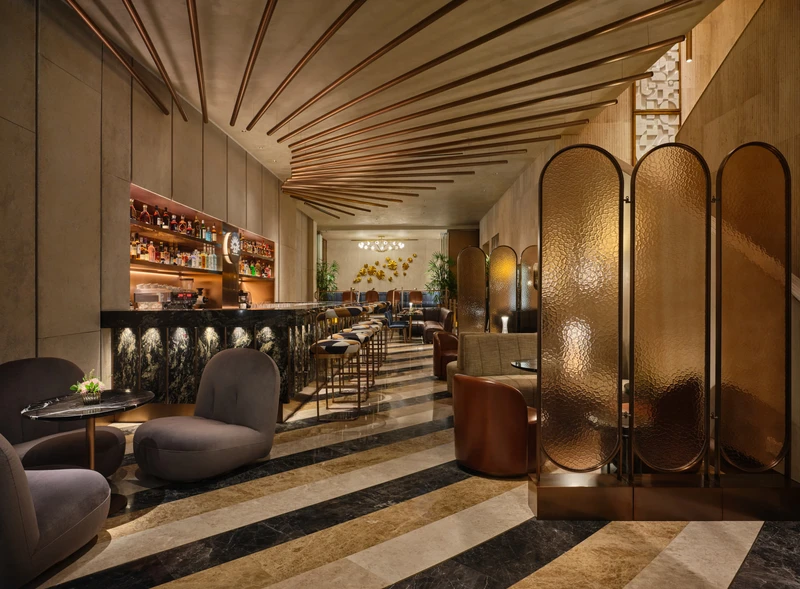 Upscale lounge bar with hammered brass folding screen, striped marble flooring, ribbed ceiling detail, velvet armchairs, and a backlit bar counter.