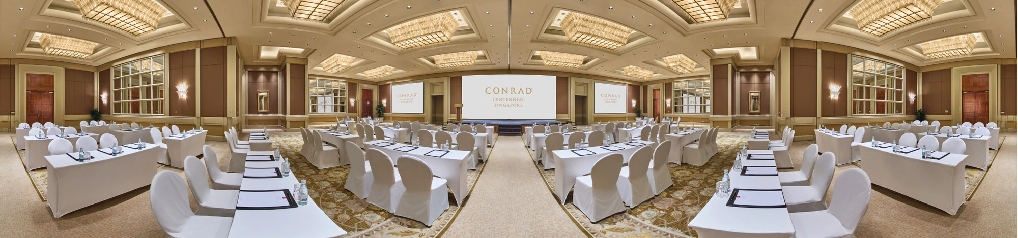 Panoramic view of a grand hotel ballroom in classroom setup with white-draped tables and chairs, central projection screen, coffered gold ceiling, and mirrored wall panels.