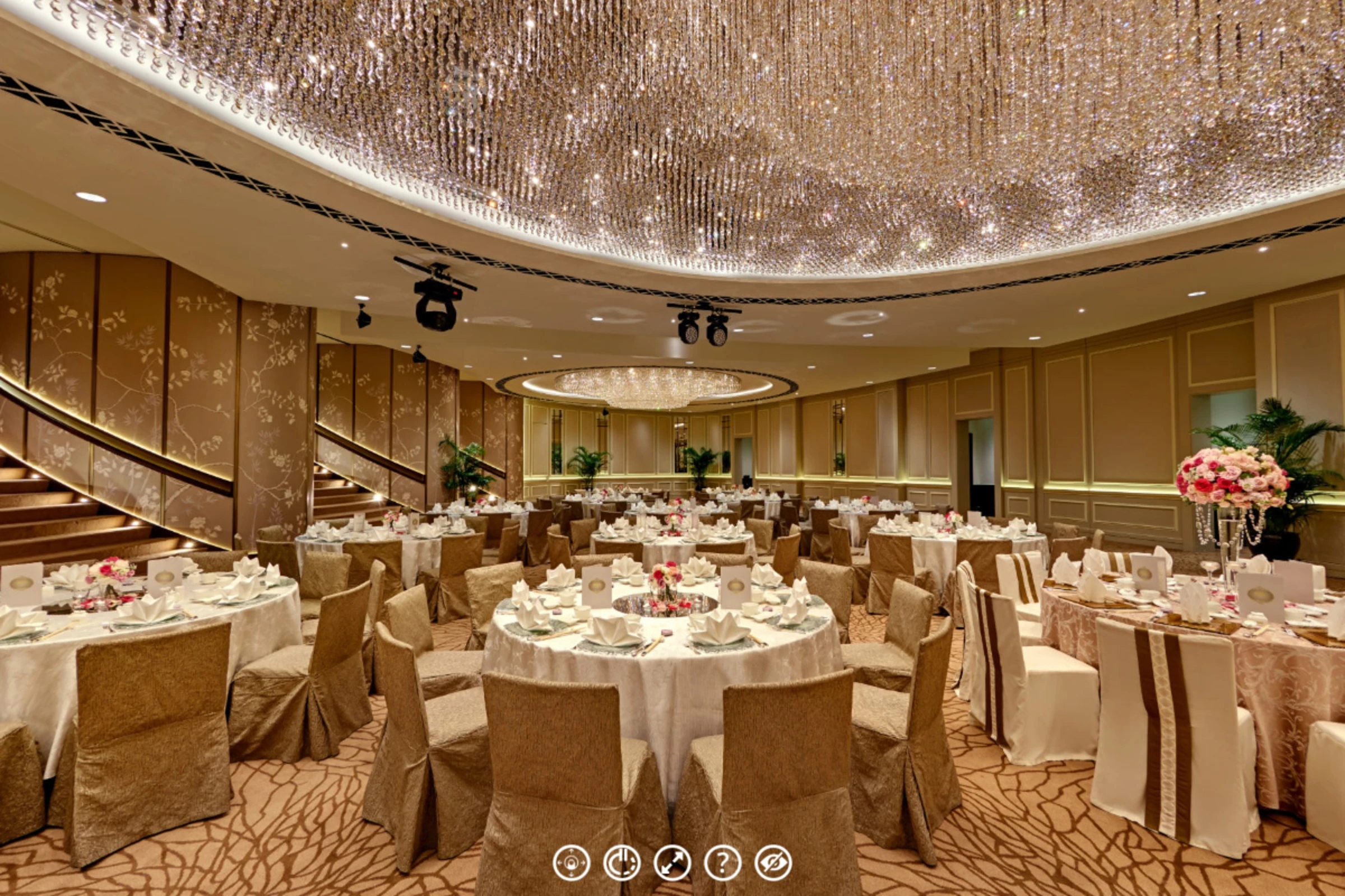 Grand ballroom with a massive crystal chandelier ceiling, round banquet tables dressed in white linen and gold chair covers, and a curved staircase at left.