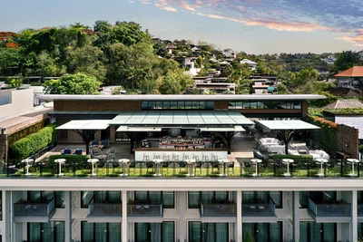 Aerial view of a rooftop bar and restaurant with open-air seating, white parasols, green lawn area, and tropical hillside villas in the background at dusk.