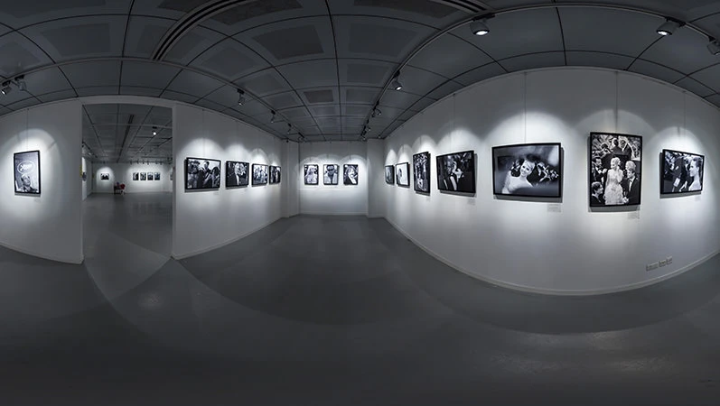 Panoramic view inside a gallery with spot-lit black-and-white photographs mounted on white walls and a dark ceiling.