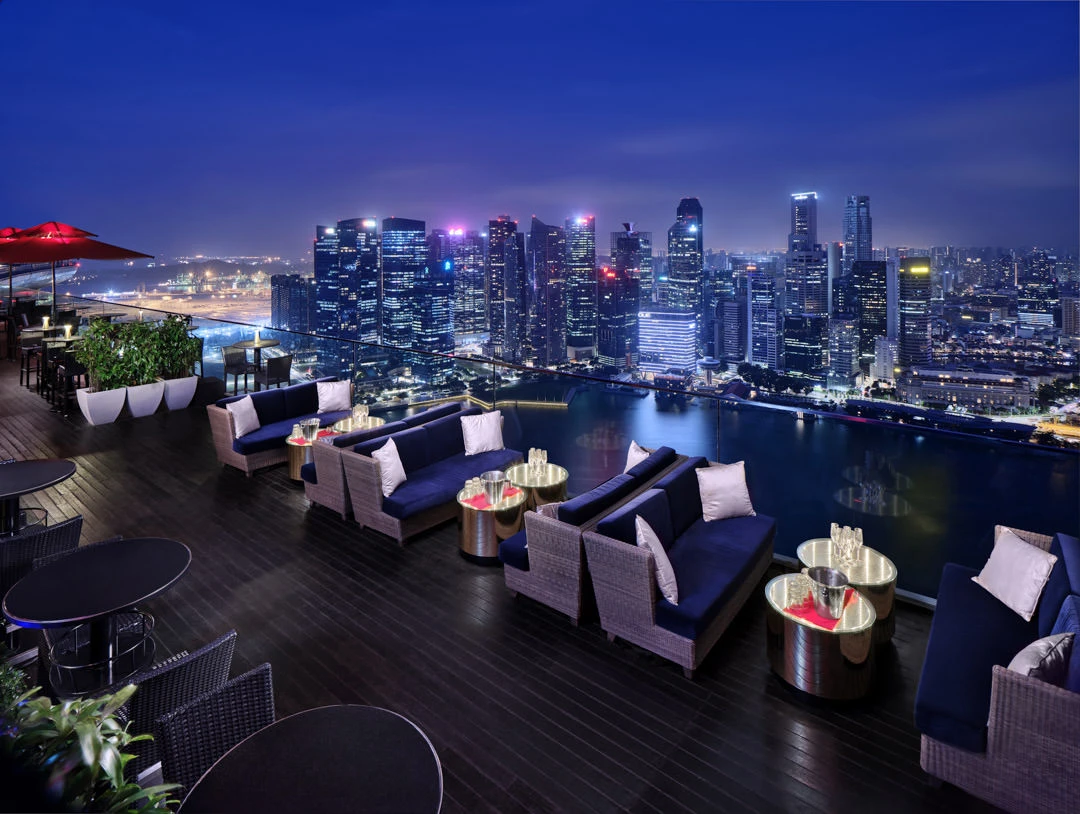 Rooftop lounge with navy cushioned seating and city skyline illuminated at dusk, dark timber decking
