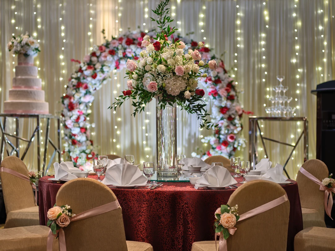 Wedding ballroom head table with tall rose and hydrangea centerpiece, burgundy damask tablecloth, floral arch backdrop, tiered cake, and fairy light curtains behind.