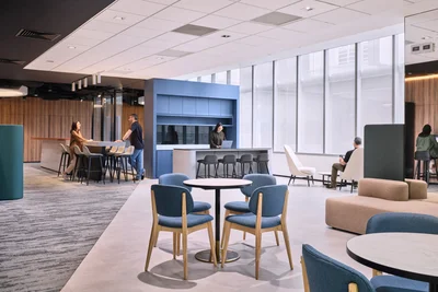 Bright open-plan office breakout area with blue chairs, kitchen counter and people mingling