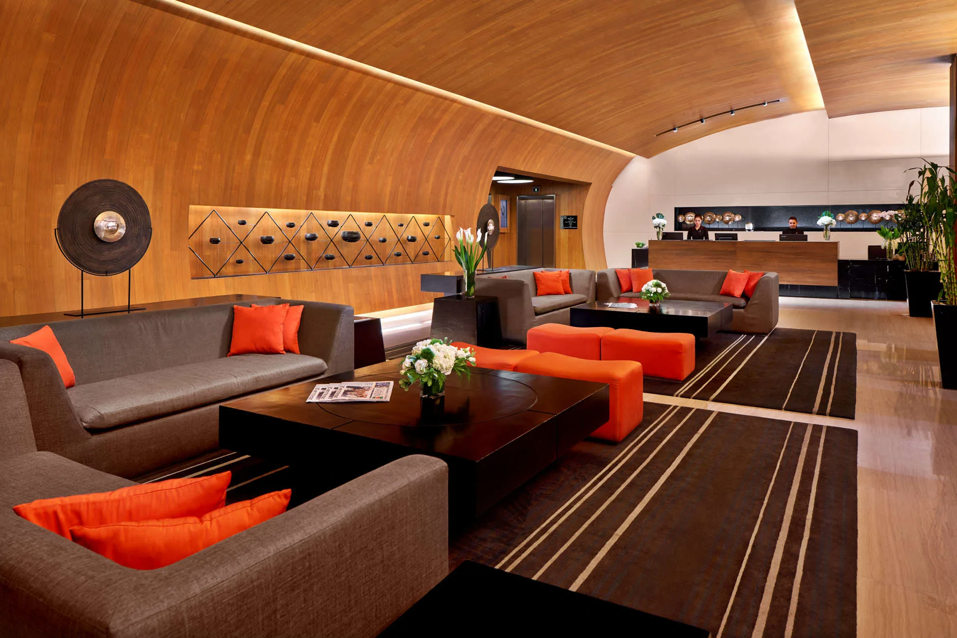Hotel lobby with a curved wooden-panelled ceiling, grey sofas with orange cushions, dark wood coffee tables, a striped rug, and a reception desk in the background.