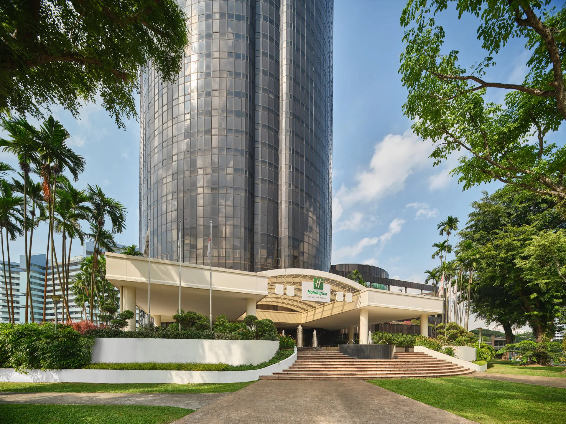 Cylindrical glass tower above landscaped entrance under blue sky — Holiday Inn Atrium Singapore architectural photography