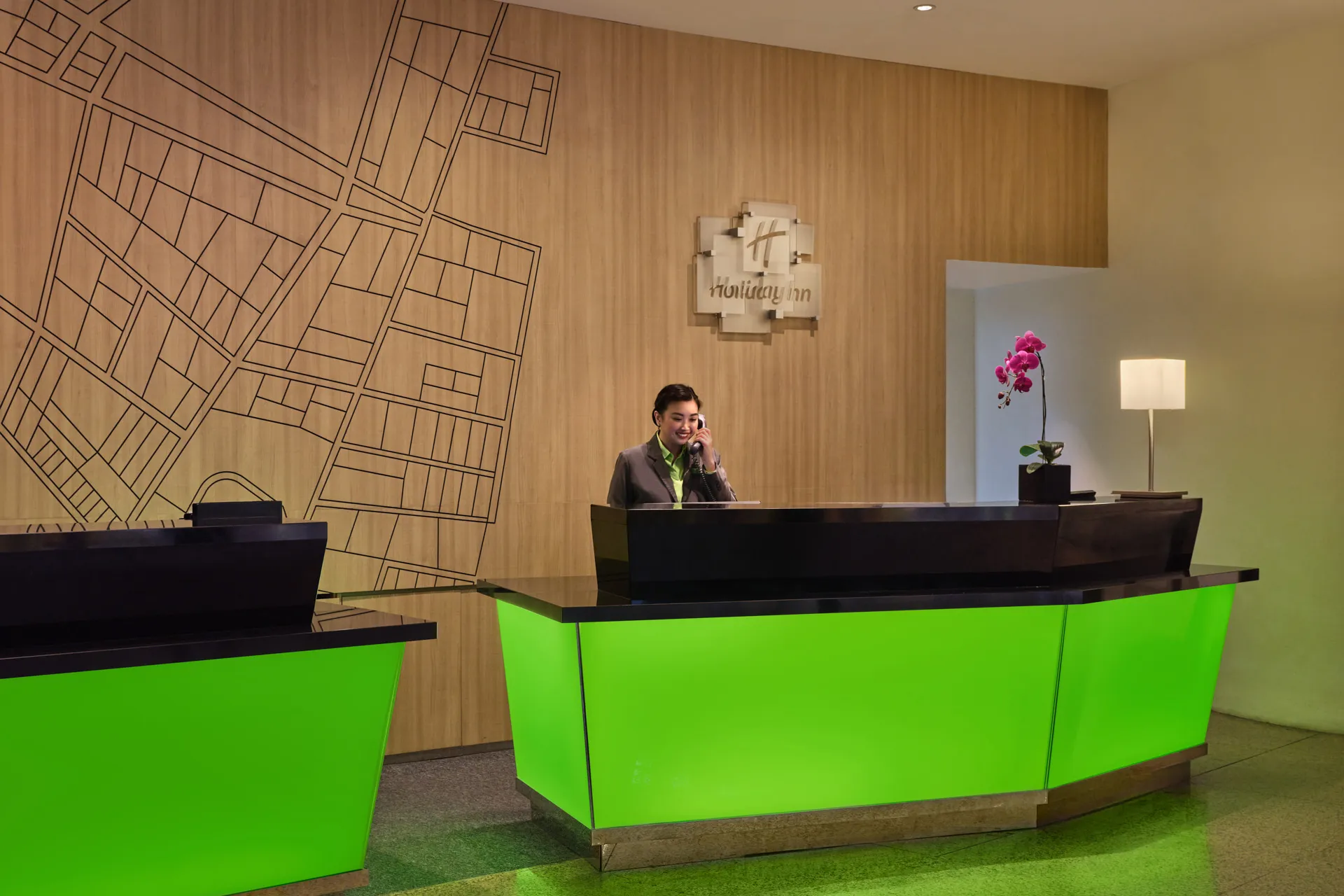 Smiling receptionist at green-lit check-in counter with wood-paneled wall — Holiday Inn Atrium Singapore hotel photography