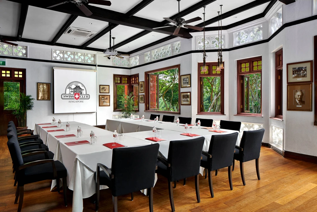Colonial-style meeting room with U-shaped white-clothed table, black leather chairs, dark timber beams, stained-glass transom windows, and a projection screen at the front.