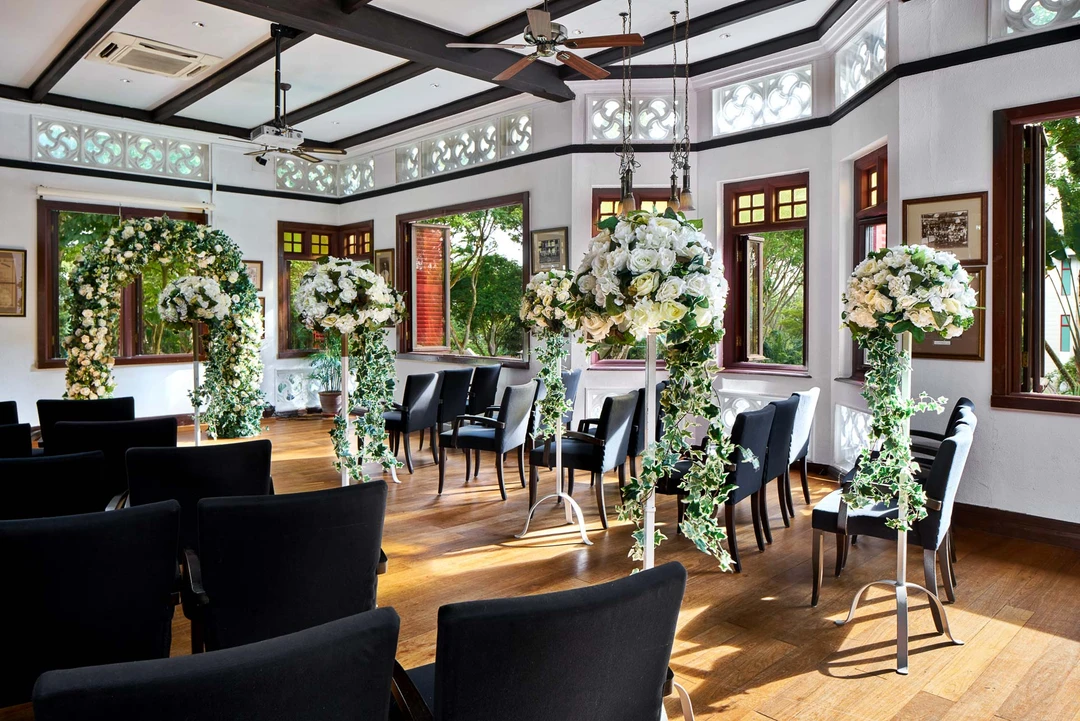 Colonial-style lounge with white walls, dark timber beams, black leather chairs arranged in rows, and tall white floral arrangements lining a central aisle.