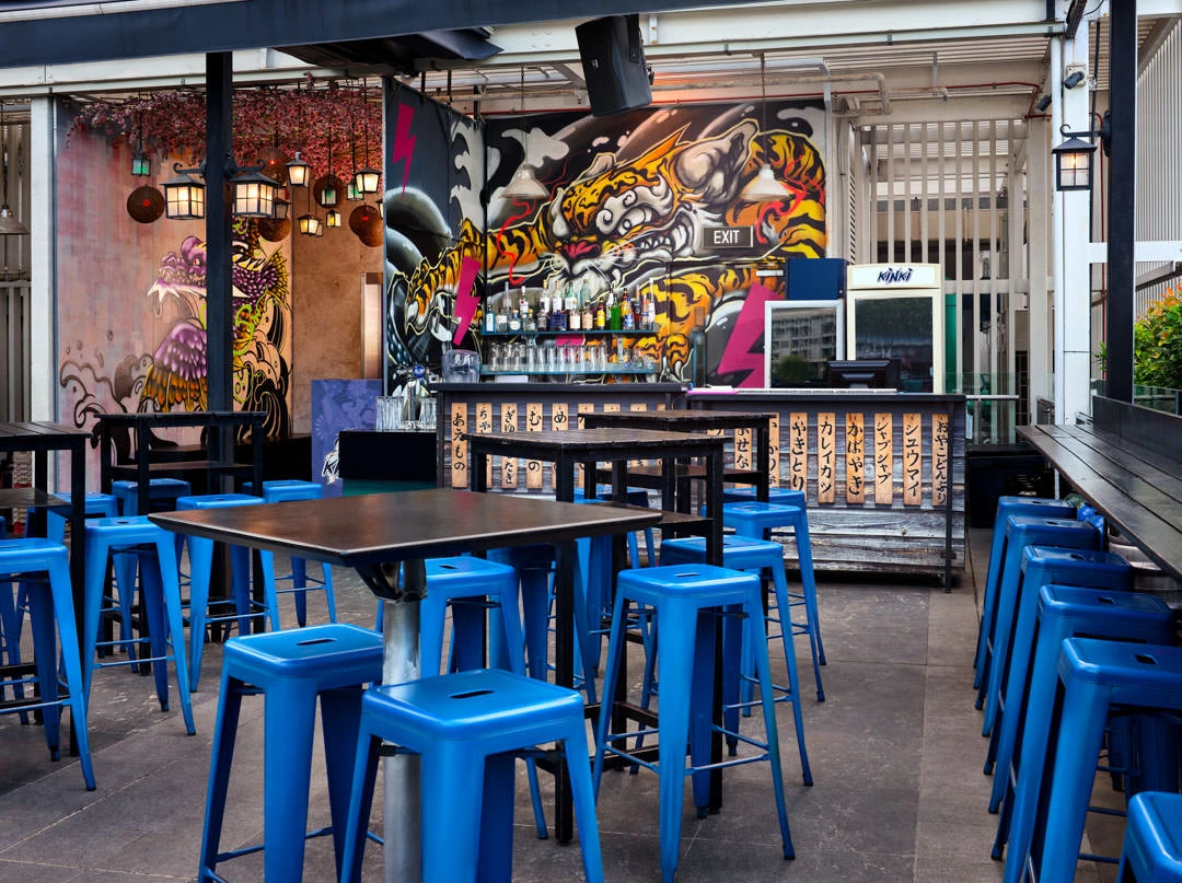 Rooftop bar area with blue metal stools around dark tables, colorful Japanese-style tiger and dragon graffiti mural behind the bar counter with lanterns.