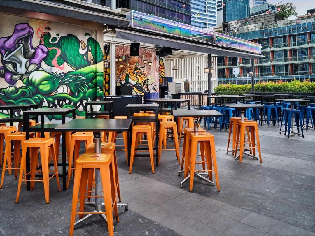 Rooftop terrace with orange and blue metal bar stools around high tables, green dragon graffiti mural on the wall, city buildings in the background.