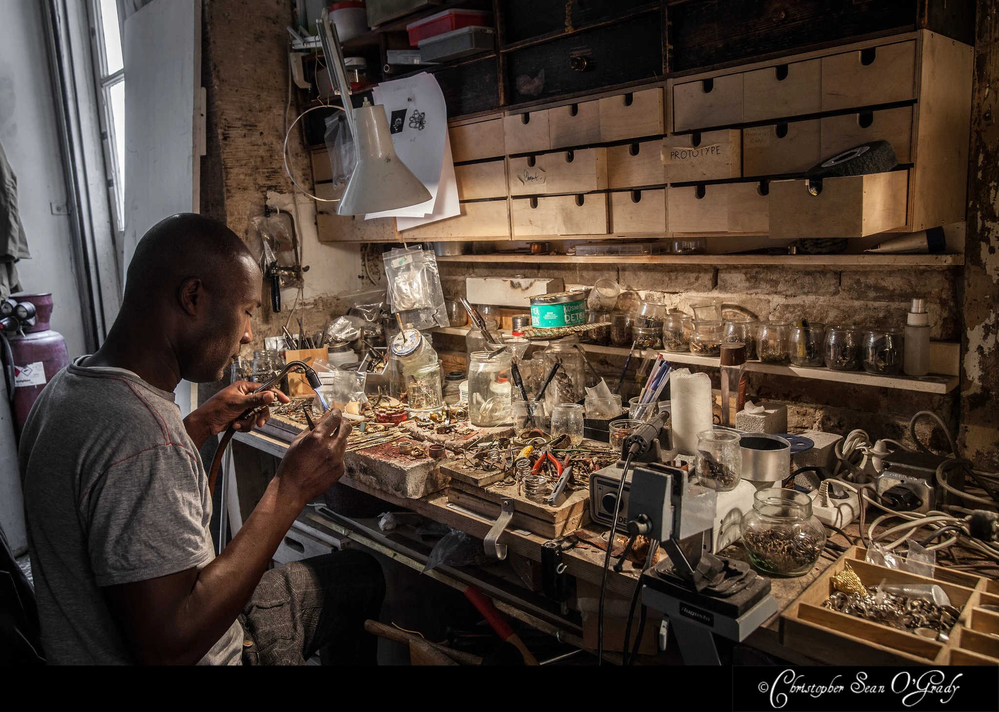 Jeweller at cluttered workbench with natural window light — Lotta Djossou Paris lifestyle portrait photography
