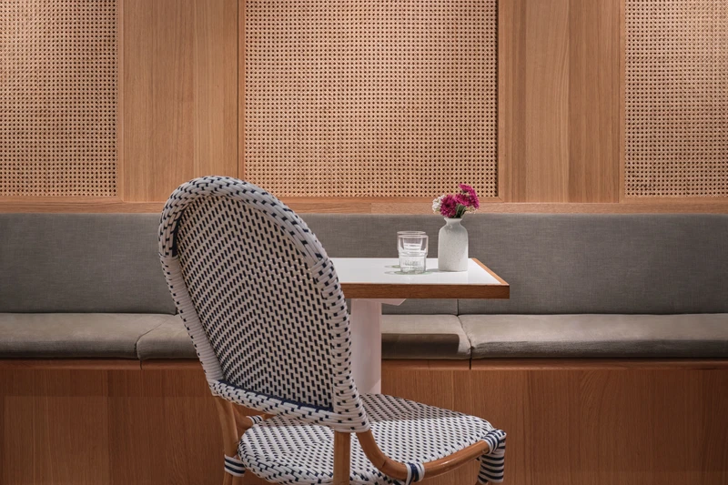 Bistro dining detail with black-and-white woven chair, grey banquette, rattan-panelled wall, water glass and flower vase on white table