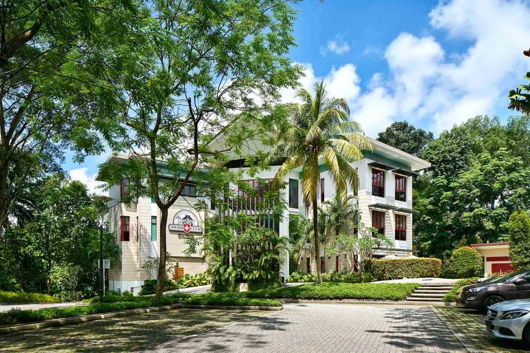 White colonial-style guesthouse with green trim, red-framed windows, surrounded by lush tropical trees and palm trees under a blue sky.