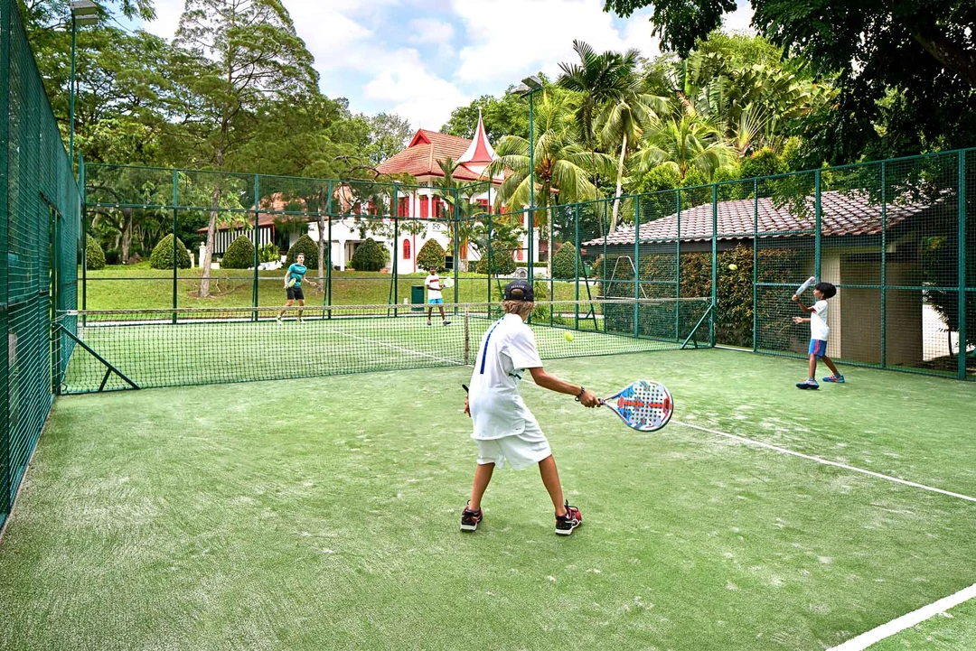 Players on a green padel court enclosed by glass and mesh walls, with tropical trees and a red-roofed clubhouse in the background.