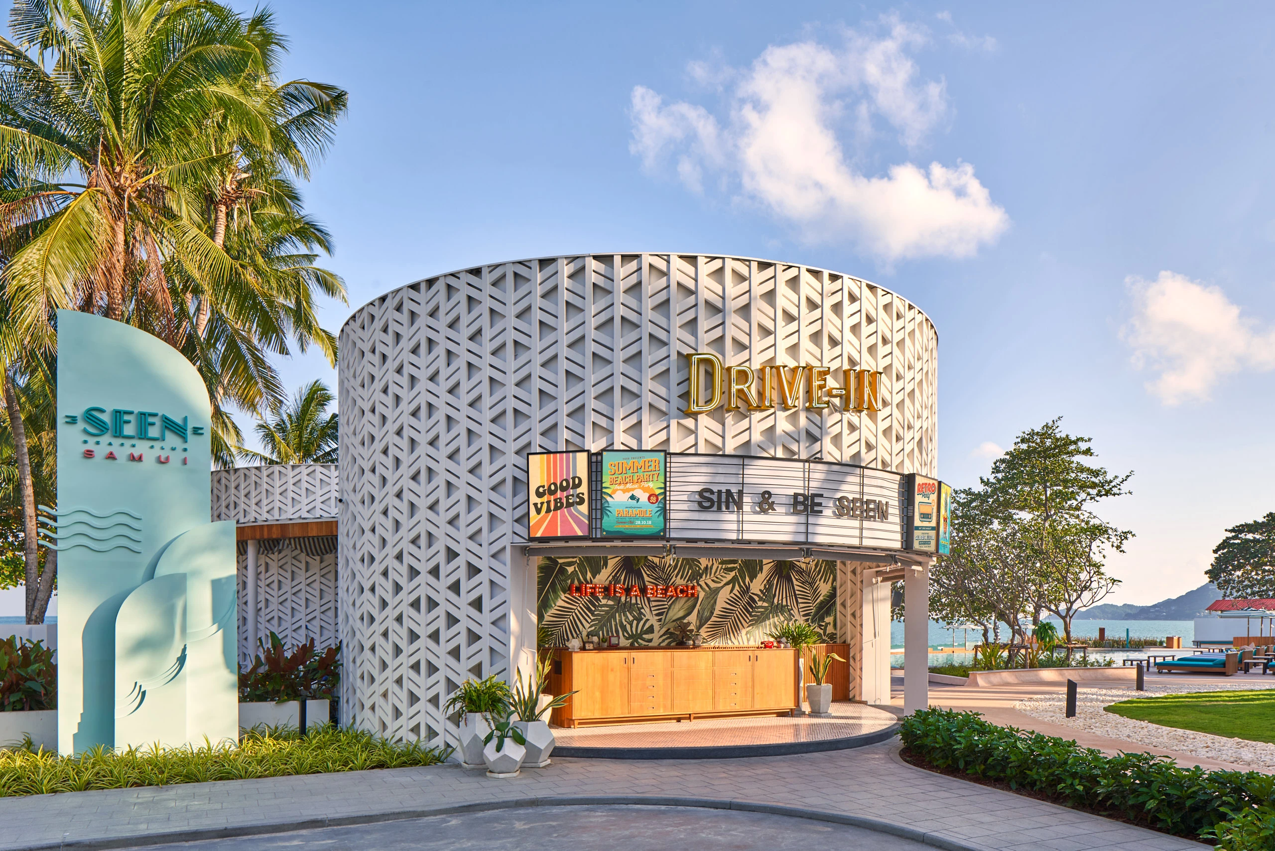 Cylindrical white lattice-screen entrance pavilion with retro drive-in marquee reading Sin and Be Seen, palm trees, and ocean views at Seen Beach Club Koh Samui