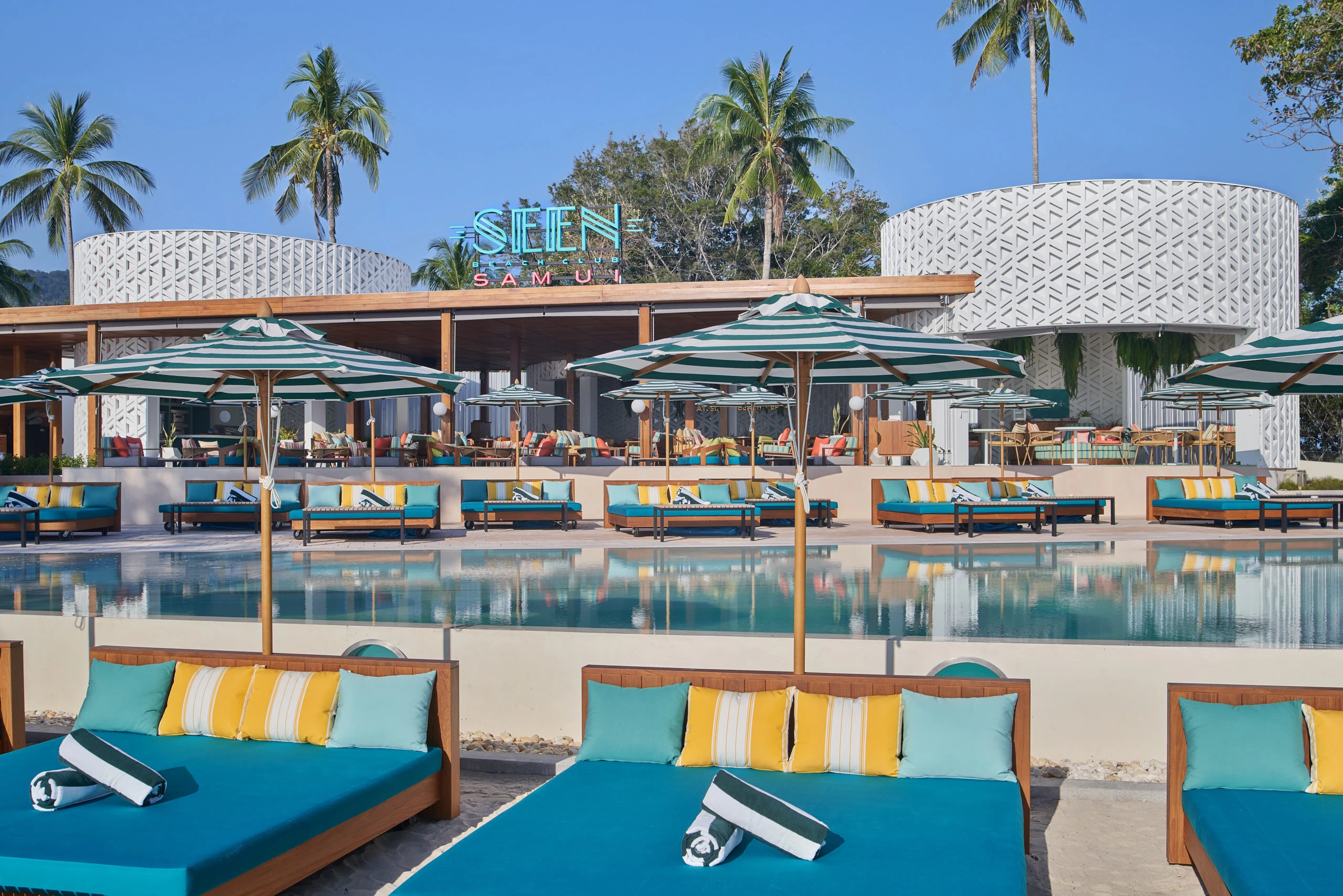 Poolside teak day beds with teal cushions and striped yellow pillows under parasols, looking toward the white lattice entrance pavilion and neon SEEN sign at Seen Beach Club Koh Samui