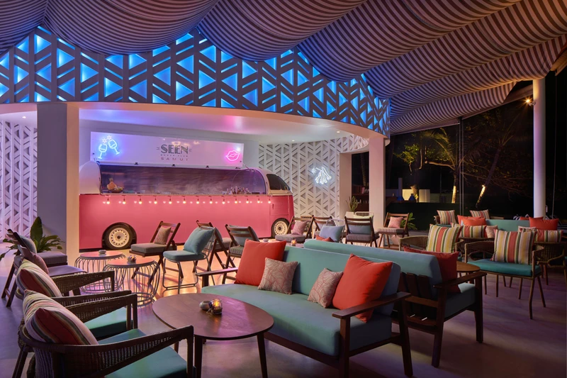 Pink vintage bus converted into a cocktail bar with neon signage under a striped canopy ceiling, blue geometric lattice screens, and teal lounge seating with colorful cushions at Seen Beach Club Koh Samui