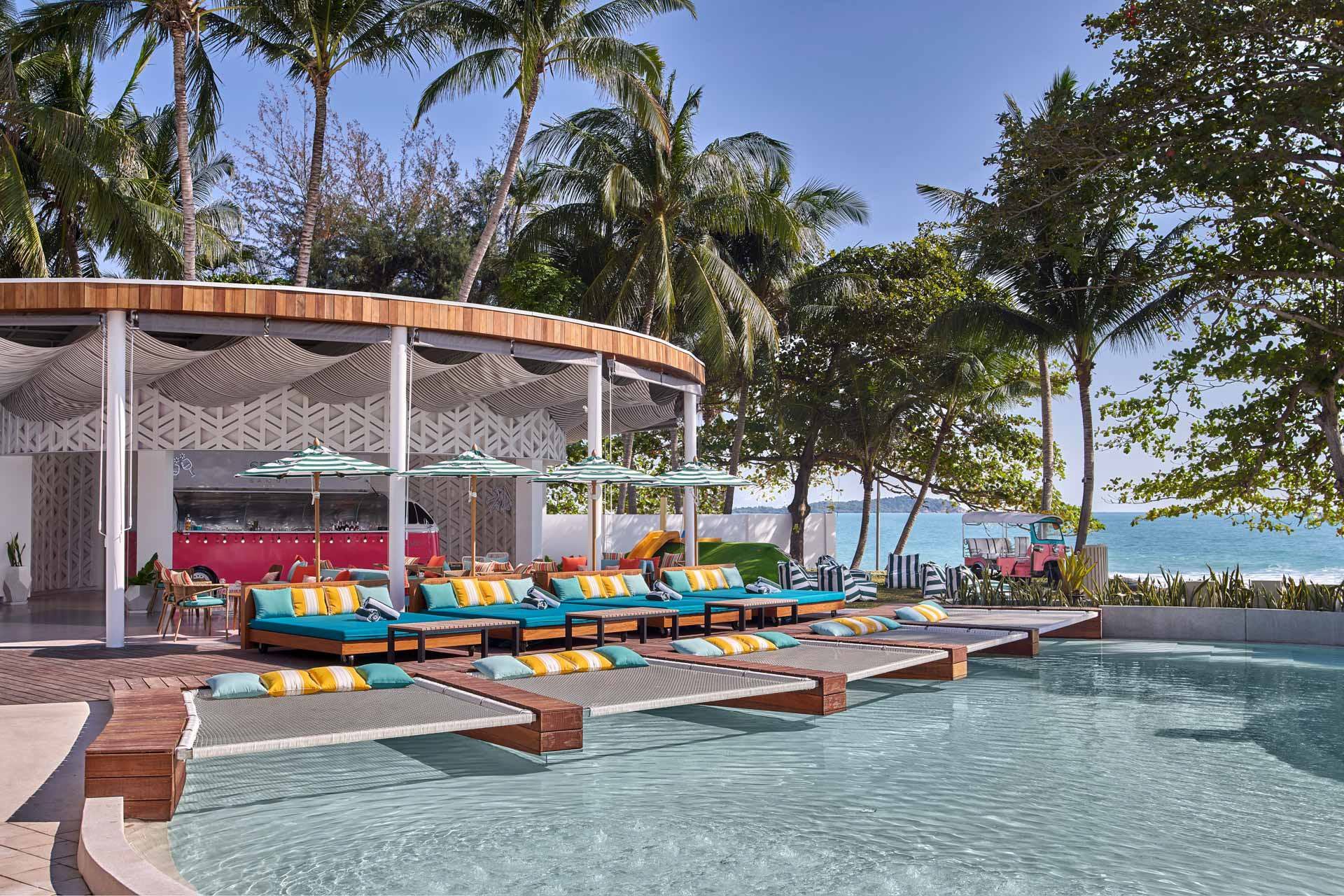 Daytime pool area with colorful cushioned day beds, curved pavilion roof, and palm trees against ocean backdrop
