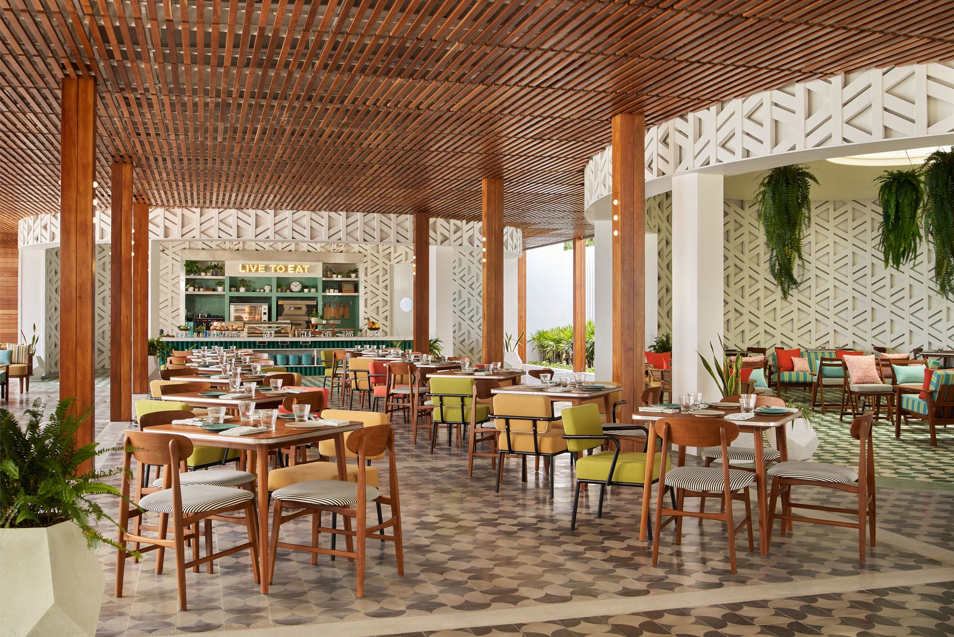 Open-air restaurant with timber-slatted ceiling, tall wooden columns, patterned tile floor, colorful mid-century chairs, and a green buffet counter at Seen Beach Club Koh Samui