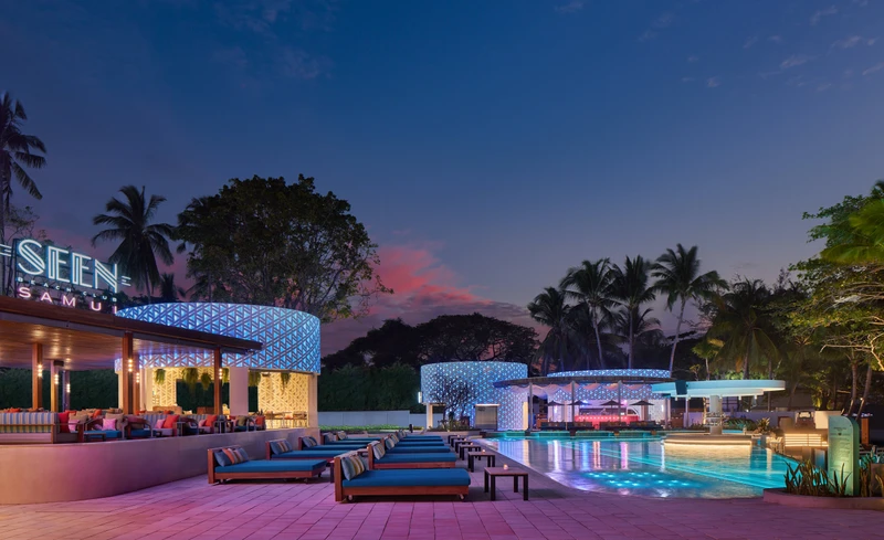 Pool terrace at sunset with blue cushioned day beds, illuminated geometric lattice pavilions glowing blue and pink, neon SEEN sign, and palm trees against a colorful dusk sky at Seen Beach Club Koh Samui