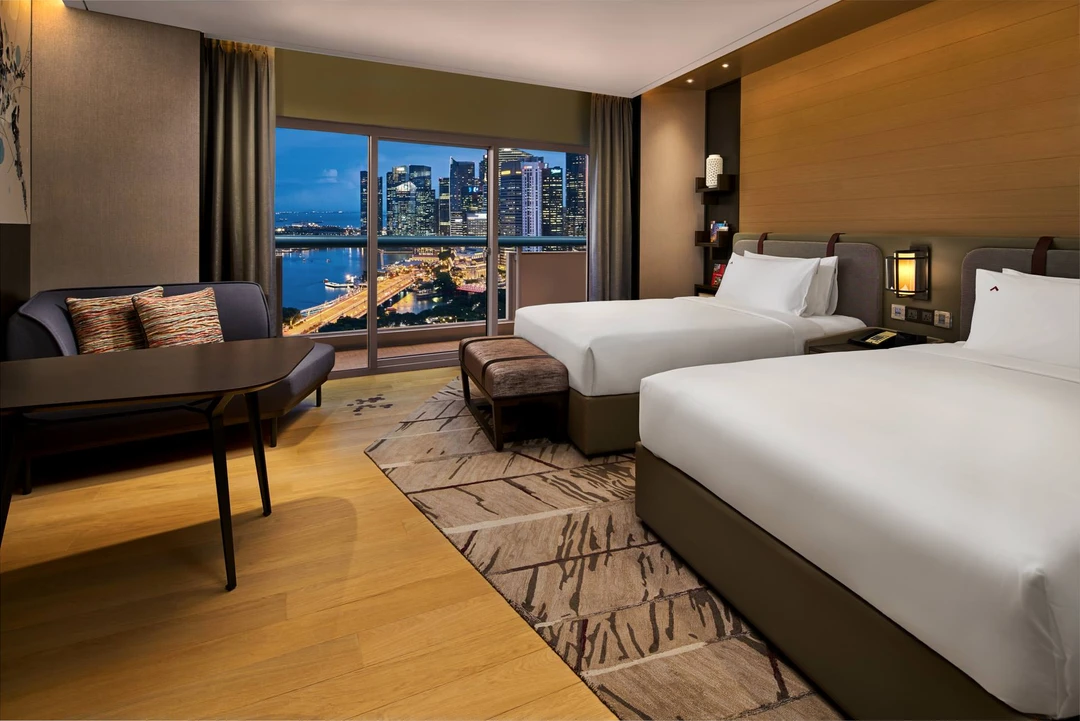 Hotel twin room with two white-sheeted beds, wood-paneled headboard, dark seating area, and harbour city views through large windows at twilight.