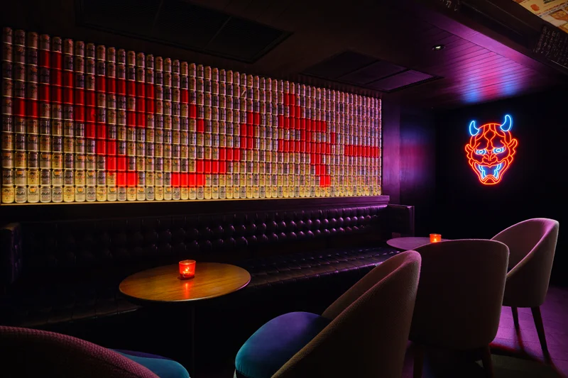 Dark bar interior with a wall of backlit sake cans forming a red pattern, a neon oni demon mask sign, tufted leather banquette, and round tables.
