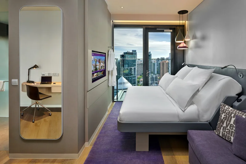 Modern hotel room with a platform bed, white duvet, purple carpet and sofa, full-length mirror, wall-mounted TV, desk area, and high-rise city views.