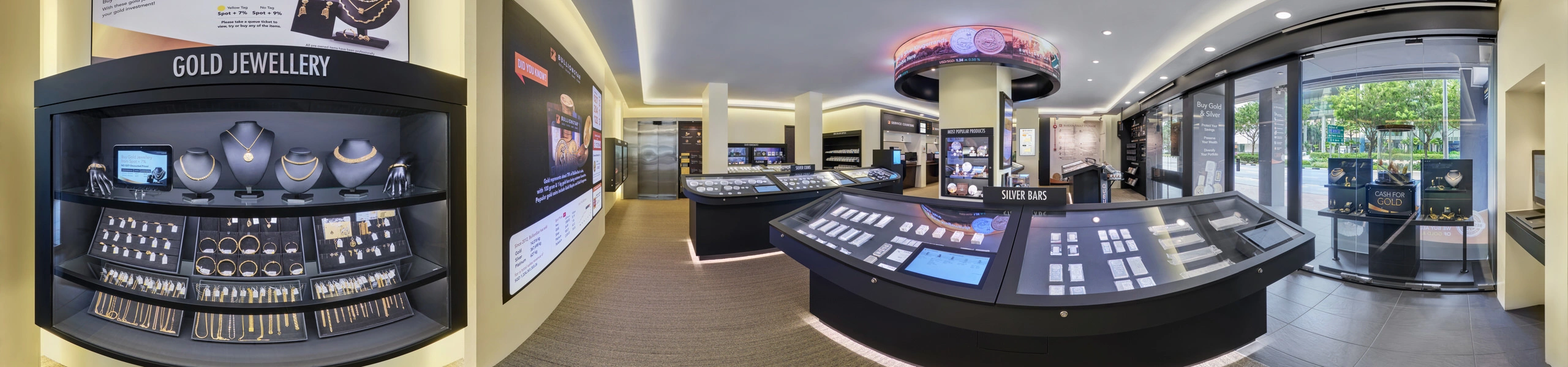 Panoramic interior of a bullion showroom with glass display cases of gold jewellery and silver bars, illuminated cabinets, a circular overhead screen, and street-facing windows.