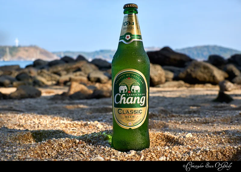 Chang Classic lager bottle on a sandy Thai beach with rocky shoreline