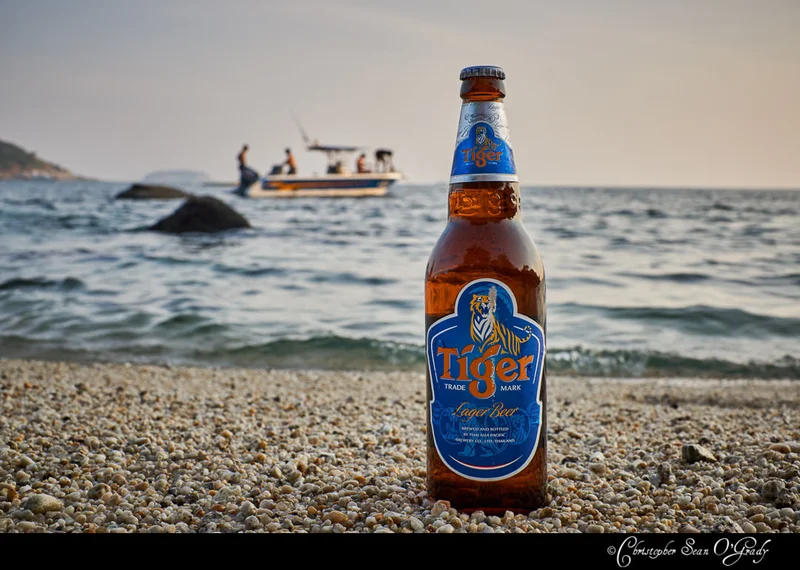 Tiger Lager Beer bottle on pebbly beach with boat on the sea