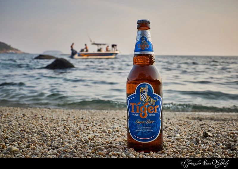 Tiger Lager Beer bottle on pebbly beach with boat on the sea