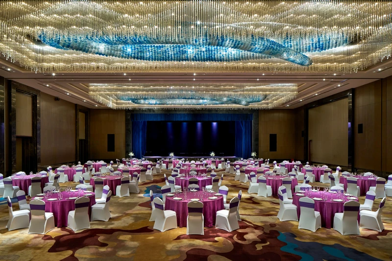 Large ballroom with round banquet tables, magenta linens, white chair covers, blue-curtained stage and crystal chandelier at Crowne Plaza Tianjin Jinnan.