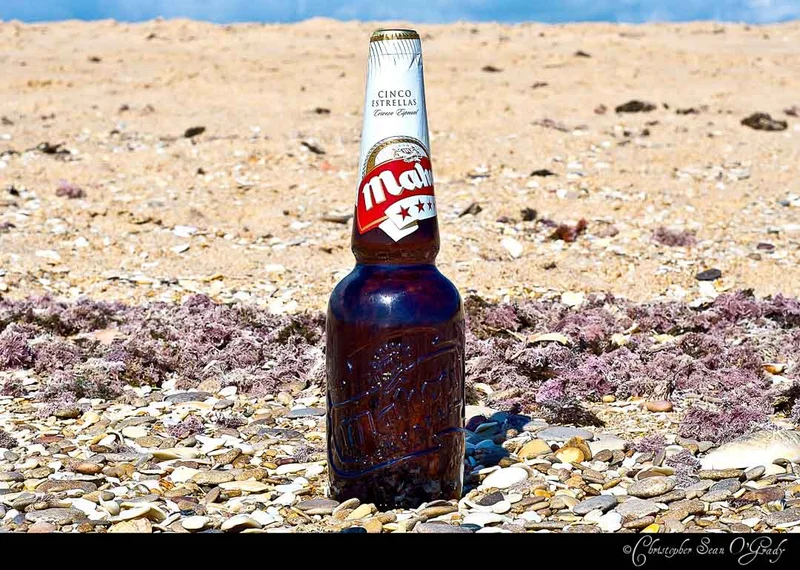 Mahou Cinco Estrellas beer bottle on a sandy beach with pebbles and seaweed