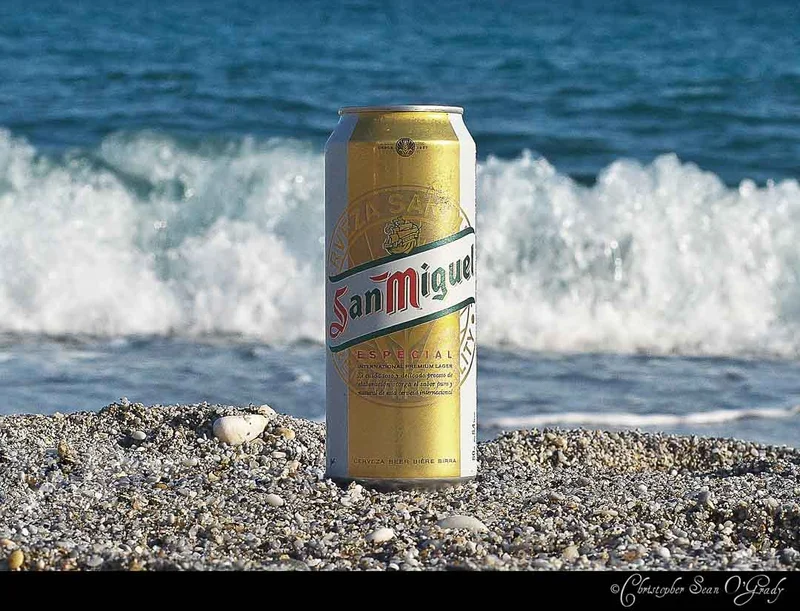 San Miguel Especial gold beer can on a pebble beach with crashing waves
