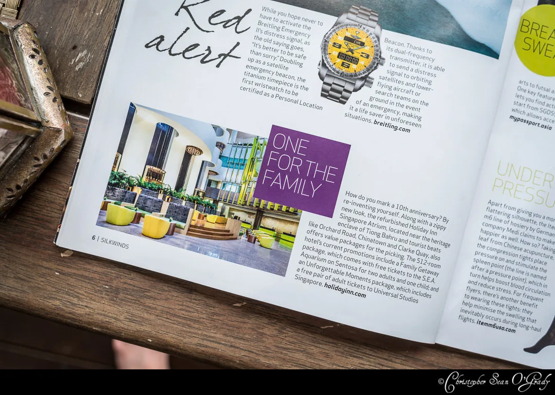 Open magazine spread on a wooden surface showing a hotel lobby photo, watch advertisement, and lifestyle articles with a watermark signature.