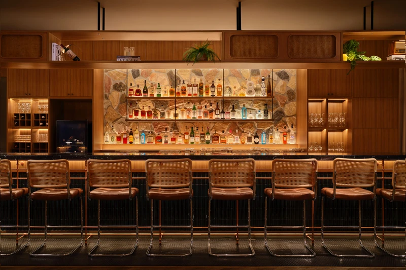 Bar counter with rattan-backed stools, backlit stone feature wall, and dark marble countertop lined with bottles