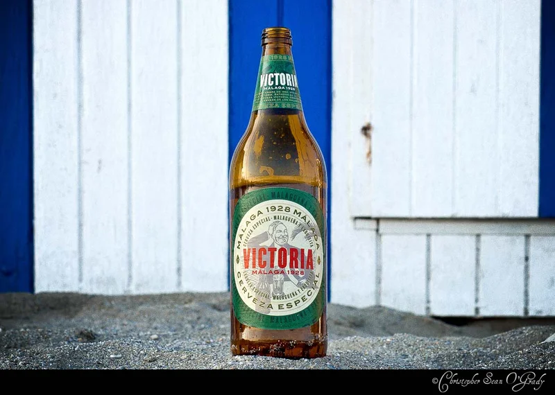 Victoria Malaga 1928 Cerveza Especial bottle on sand by a blue and white beach hut