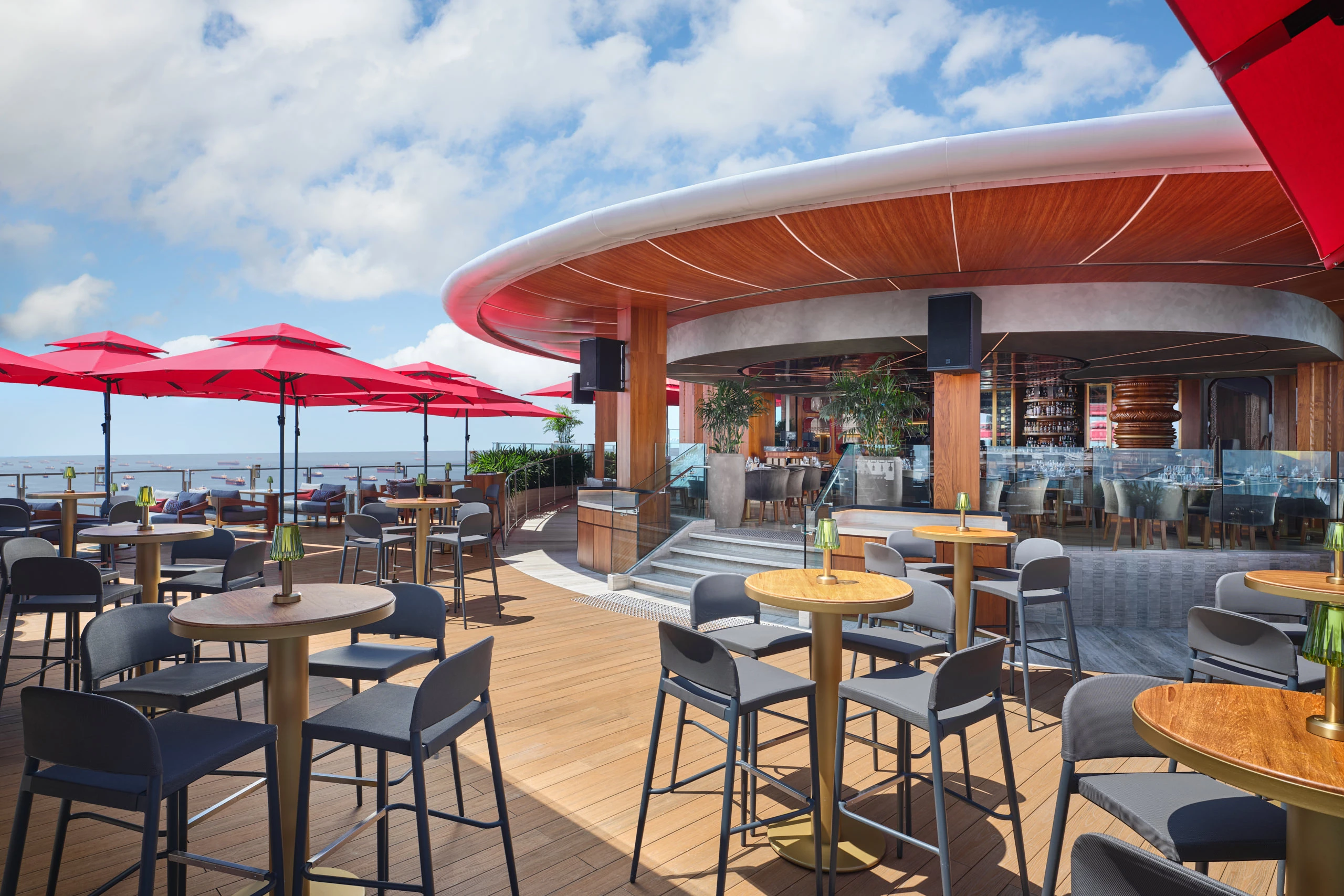 Rooftop restaurant terrace with high tables and grey stools, red parasols, curved wooden-clad canopy overhead, glass-walled bar area, and open sky beyond.