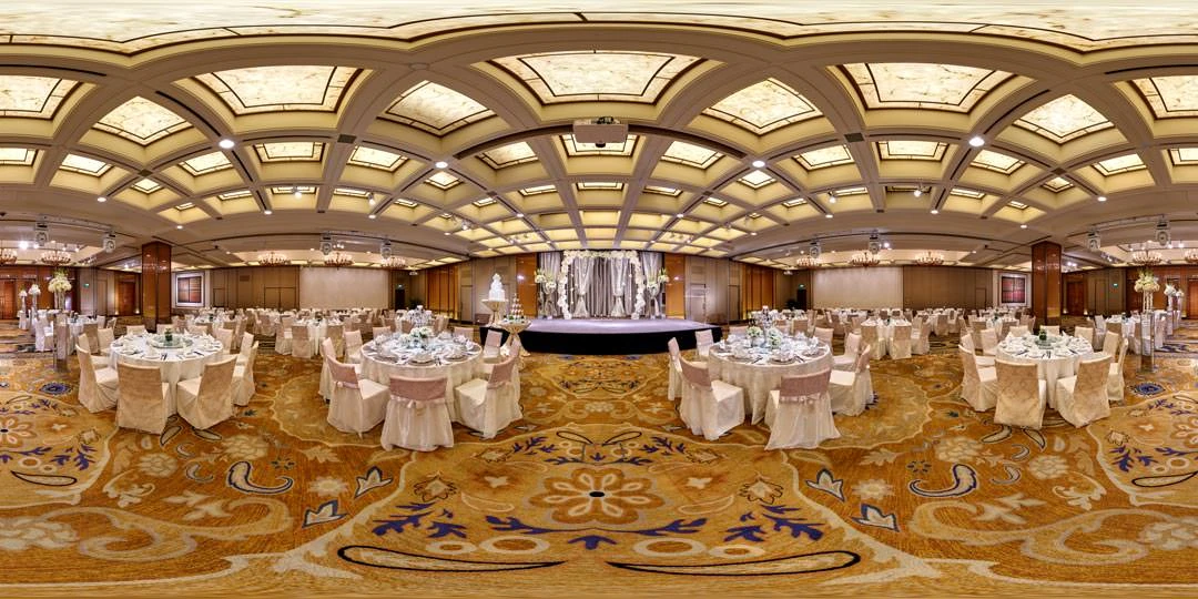 Grand ballroom set for a wedding banquet with round tables in ivory linens, tall floral centrepieces, a stage with draped backdrop, and arched golden ceiling panels.