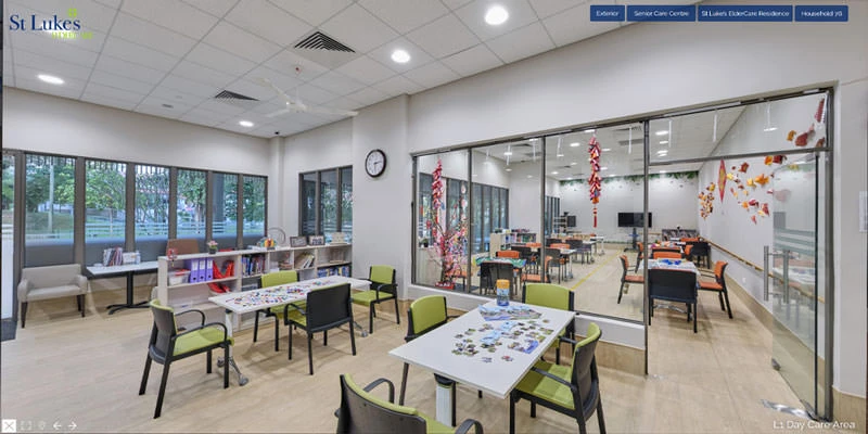 Bright eldercare day-care room with activity tables, green-and-grey chairs, puzzle materials, glass partition walls, and colourful paper decorations hanging from the ceiling.