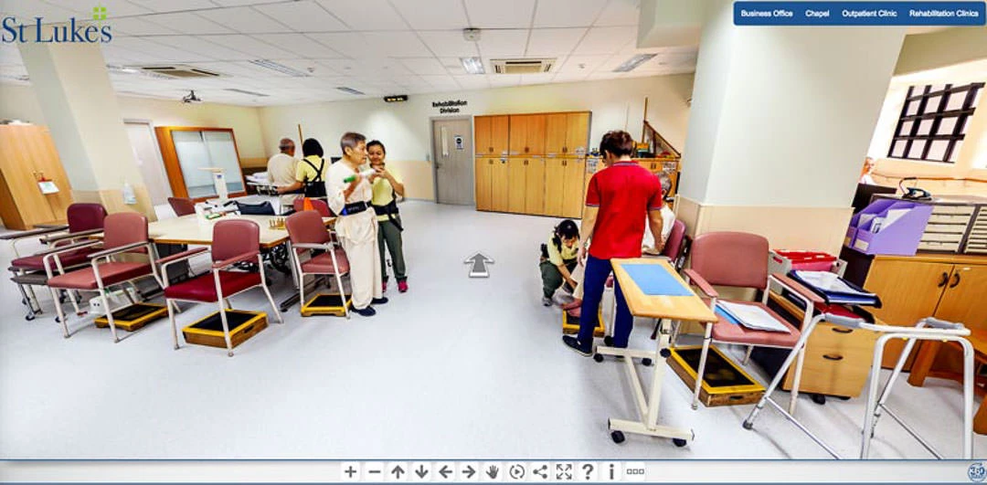 Hospital rehabilitation room with staff assisting a patient in a walking harness, surrounded by therapy tables, red chairs, and mobility aids.