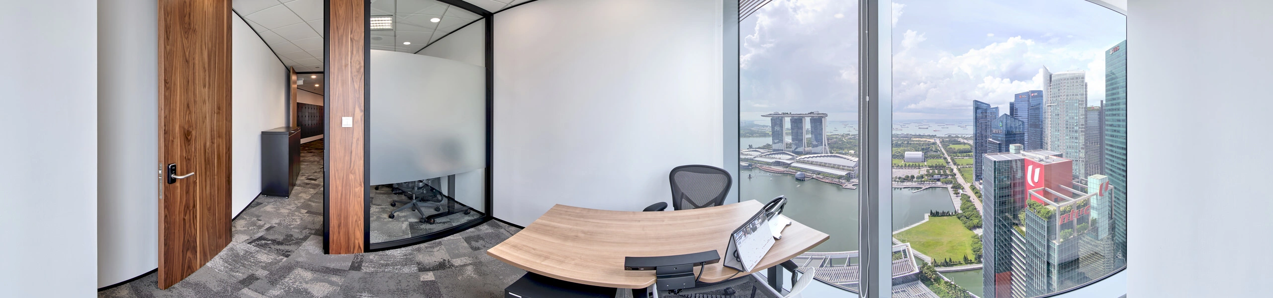 Panoramic view of a private office with a curved wooden desk, mesh chair, and floor-to-ceiling windows overlooking Marina Bay Sands and the Singapore skyline.