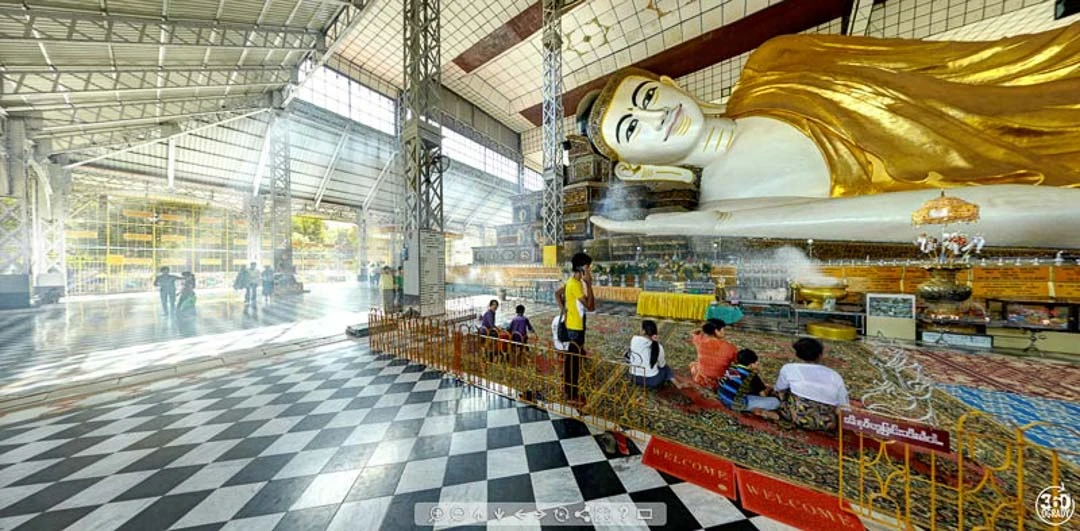 Massive reclining Buddha statue in gold and white inside a covered pavilion with black-and-white chequered floor and visitors seated nearby.