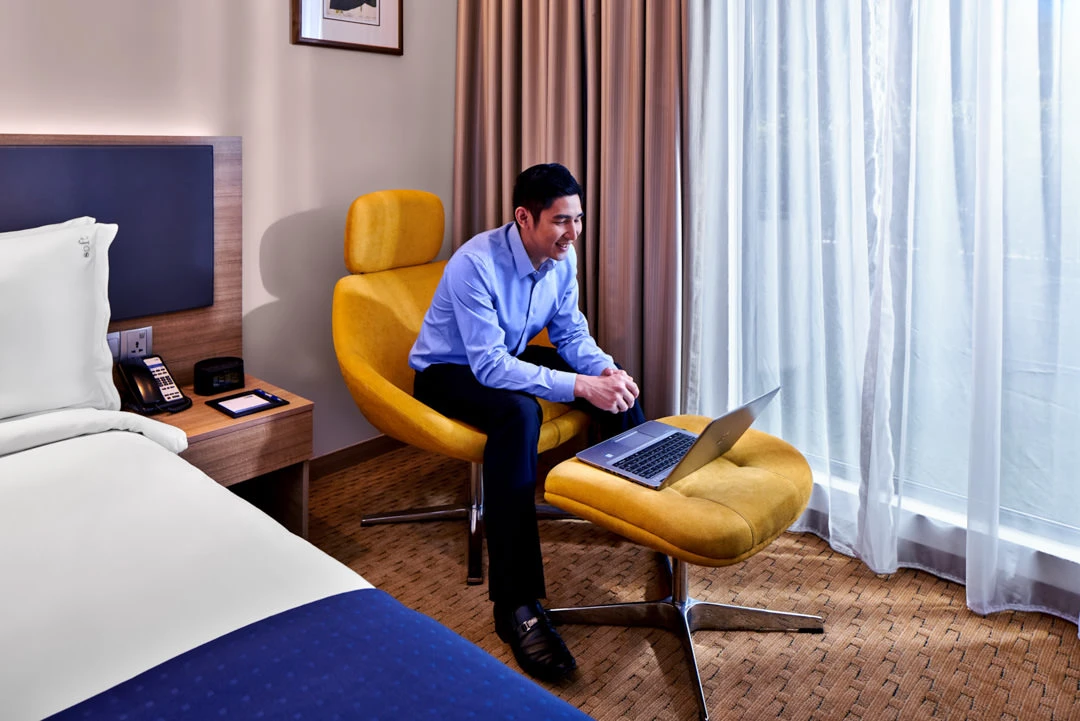 Guest with laptop in mustard lounge chair beside bed