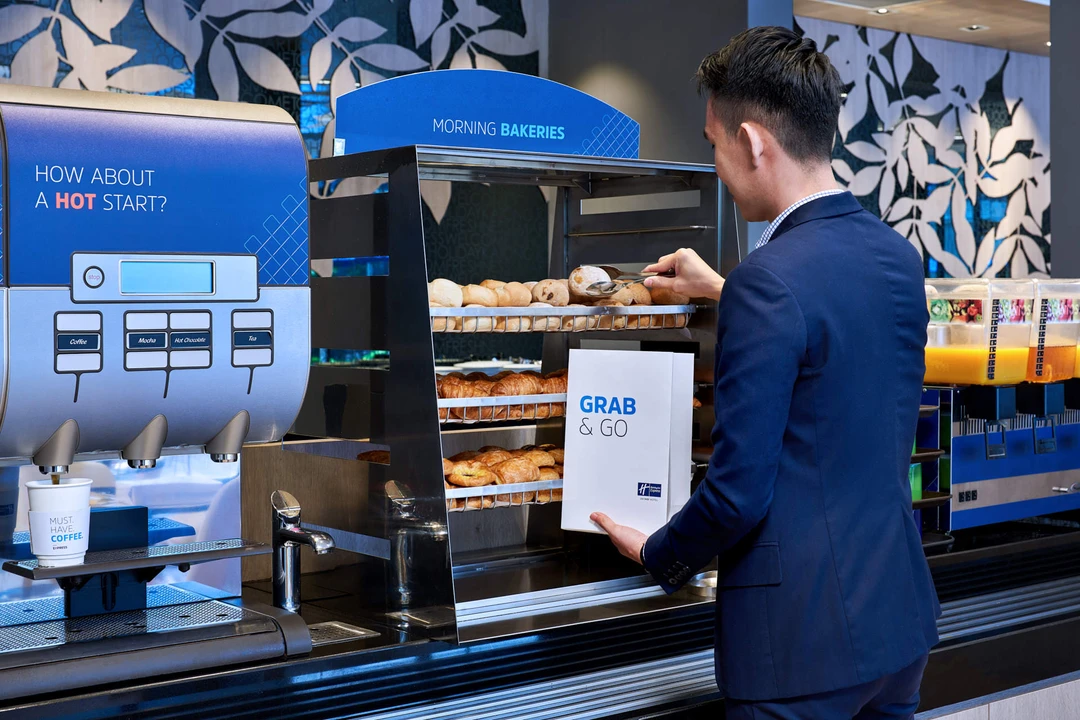 Man in dark shirt selecting pastries from a tiered bakery display at a hotel breakfast station with coffee machine and orange juice dispensers nearby.
