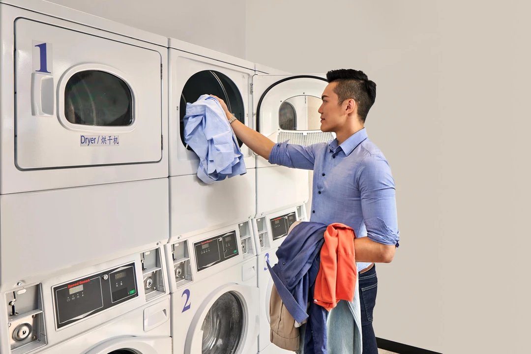 Man in blue shirt loading colorful clothing into a stacked white commercial dryer in a hotel self-service laundromat with numbered machines.