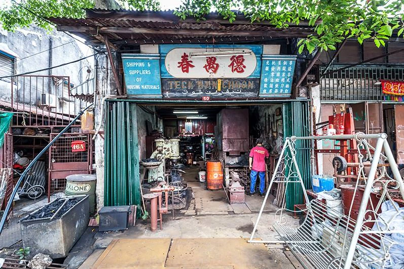 Weathered shophouse foundry entrance with Chinese signage, green metal doors, machinery and metal parts scattered outside, and a worker inside.